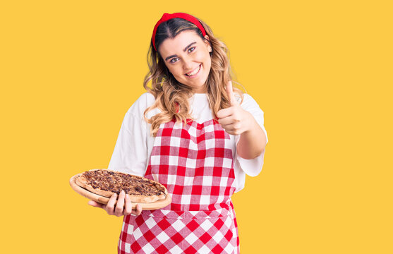Young caucasian woman wearing apron holding pizza smiling happy and positive, thumb up doing excellent and approval sign