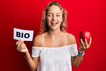 Beautiful caucasian woman holding red apple and bio word winking looking at the camera with sexy...