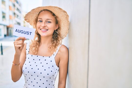 Young beautiful caucasian woman with blond hair smiling happy outdoors on a summer day holding August word