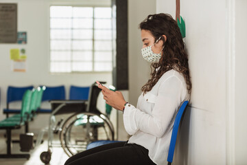 woman in a mask sitting on a hospital bench waiting to be seen, fiddling with her cell phone
