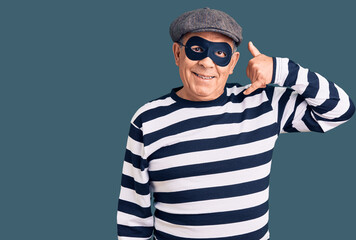 Senior handsome man wearing burglar mask and t-shirt smiling doing phone gesture with hand and fingers like talking on the telephone. communicating concepts.