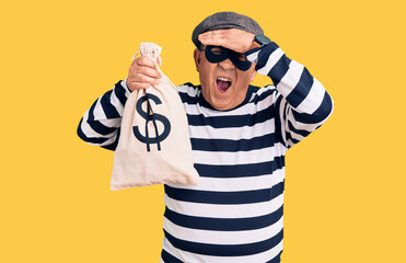 Senior handsome man wearing burglar mask holding money bag stressed and frustrated with hand on head, surprised and angry face