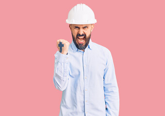 Young handsome man wearing architect hardhat angry and mad raising fist frustrated and furious while shouting with anger. rage and aggressive concept.