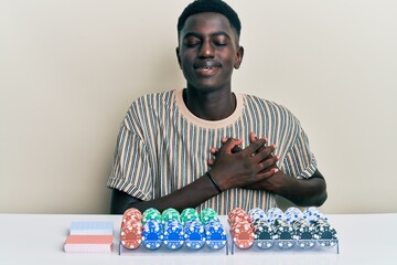 Young african american man sitting on the table with poker chips and cards smiling with hands on chest with closed eyes and grateful gesture on face. health concept.