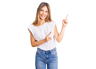 Beautiful caucasian woman with blonde hair wearing casual white tshirt smiling and looking at the camera pointing with two hands and fingers to the side.