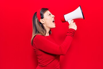 Young beautiful woman wearing diadem with angry expression. Screaming loud using megaphone standing over isolated red background