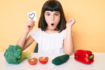 Young little girl with bang sitting on the table with veggies holding heart reminder scared and amazed with open mouth for surprise, disbelief face