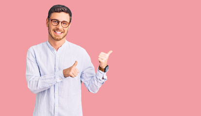 Handsome young man with bear wearing elegant business shirt and glasses pointing to the back behind with hand and thumbs up, smiling confident