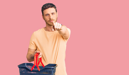 Handsome young man with bear holding supermarket shopping basket pointing with finger to the camera and to you, confident gesture looking serious