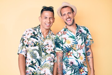 Young gay couple of two men wearing summer hat and hawaiian shirt winking looking at the camera...