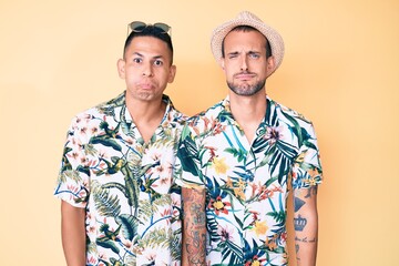 Young gay couple of two men wearing summer hat and hawaiian shirt puffing cheeks with funny face. mouth inflated with air, crazy expression.