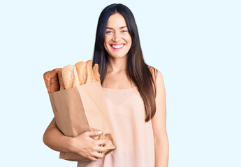 Young beautiful caucasian woman holding paper bag with bread looking positive and happy standing and smiling with a confident smile showing teeth