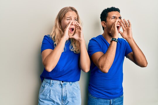 Young interracial couple wearing casual clothes shouting angry out loud with hands over mouth
