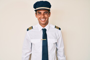 Young hispanic man wearing airplane pilot uniform winking looking at the camera with sexy expression, cheerful and happy face.