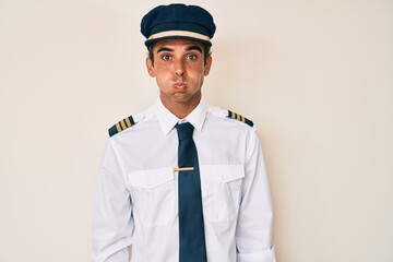 Young hispanic man wearing airplane pilot uniform puffing cheeks with funny face. mouth inflated with air, crazy expression.