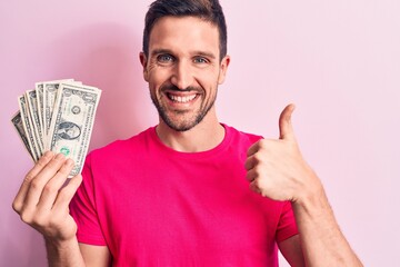 Young handsome man holding bunch of dollars banknotes over isolated pink background smiling happy and positive, thumb up doing excellent and approval sign