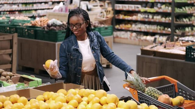 Black young woman is chosing an orange. She is doing shopping in a supermarket. The closeup view.