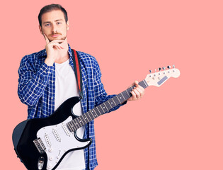 Young handsome caucasian man playing electric guitar serious face thinking about question with hand...