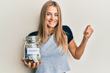 Obraz premium Young blonde woman holding charity jar with money screaming proud, celebrating victory and success very excited with raised arm