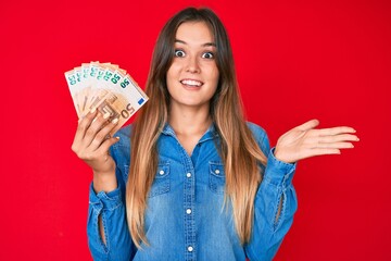 Beautiful caucasian woman holding 50 euro banknotes celebrating achievement with happy smile and winner expression with raised hand