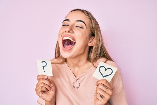 Young beautiful blonde woman holding heart and question mark reminder smiling and laughing hard out loud because funny crazy joke.