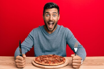 Handsome hispanic man eating tasty pepperoni pizza celebrating crazy and amazed for success with open eyes screaming excited.