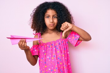 African american child with curly hair holding paper airplane with angry face, negative sign...