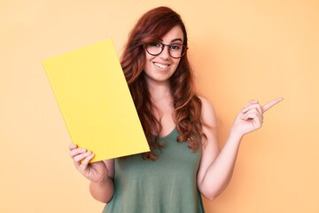 Young beautiful woman wearing glasses holding book smiling happy pointing with hand and finger to the side