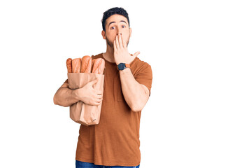 Naklejka premium Young hispanic man holding paper bag with bread covering mouth with hand, shocked and afraid for mistake. surprised expression