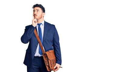 Young hispanic man wearing suit and leather bag serious face thinking about question with hand on chin, thoughtful about confusing idea