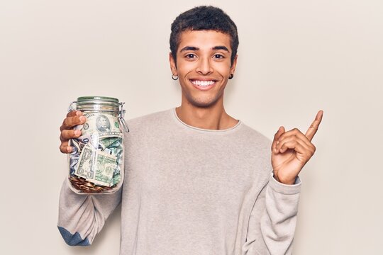 Young african amercian man holding jar with savings smiling happy pointing with hand and finger to the side