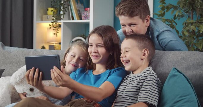 Four overjoyed children in domestic outfit looking at tablet screen and smiling while sitting on grey couch. Concept of friendship, entertainment and technology.