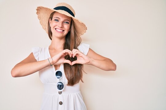 Young blonde girl wearing summer hat smiling in love doing heart symbol shape with hands. romantic concept.