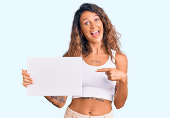 Young hispanic woman with tattoo holding blank empty paper smiling happy pointing with hand and...