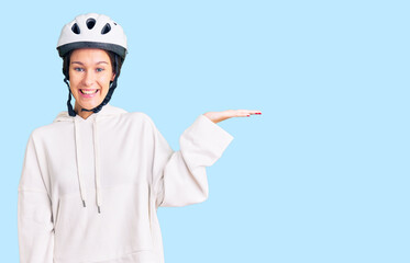Beautiful brunette young woman wearing bike helmet and sporty clothes smiling cheerful presenting and pointing with palm of hand looking at the camera.