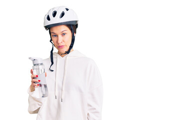Beautiful brunette young woman wearing bike helmet and holding water bottle thinking attitude and sober expression looking self confident