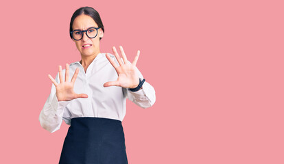 Beautiful brunette young woman wearing professional waitress apron afraid and terrified with fear expression stop gesture with hands, shouting in shock. panic concept.