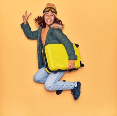 Young beautiful latin woman wearing winter clothes smiling happy. Jumping with smile on face holding cabin bag doing victory sign over isolated yellow background