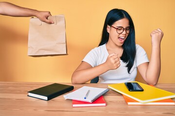 Naklejka premium Young beautiful asian girl sitting on the table stuying getting take away food celebrating surprised and amazed for success with arms raised and eyes closed