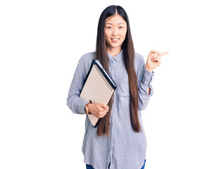 Young beautiful chinese woman holding notebook smiling happy pointing with hand and finger to the side