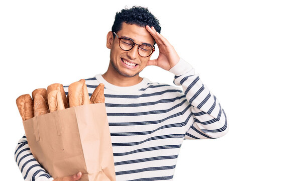 Hispanic handsome young man holding paper bag with bread stressed and frustrated with hand on head, surprised and angry face