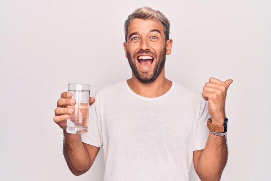 Handsome blond man with beard drinking glass of water to refreshment over white background pointing thumb up to the side smiling happy with open mouth