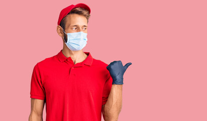 Handsome blond man with beard wearing delivery uniform and medical mask smiling with happy face looking and pointing to the side with thumb up.