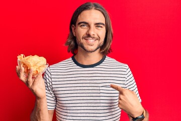 Young handsome man holding potato chip pointing finger to one self smiling happy and proud