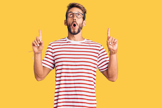 Handsome blond man with beard wearing casual clothes and glasses amazed and surprised looking up and pointing with fingers and raised arms.