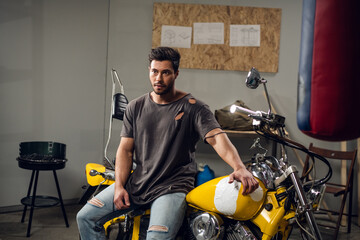 In the garage, a car mechanic sits on a yellow motorcycle that he has repaired. Pumped up stylish...