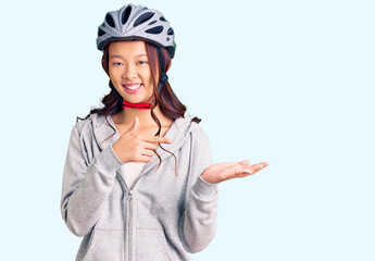 Young beautiful chinese girl wearing bike helmet amazed and smiling to the camera while presenting with hand and pointing with finger.
