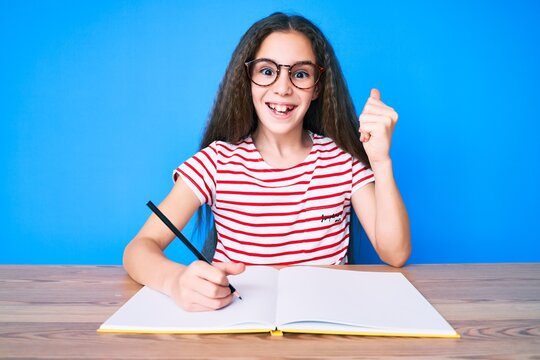Cute hispanic child girl sitting on the table writing book screaming proud, celebrating victory and success very excited with raised arms