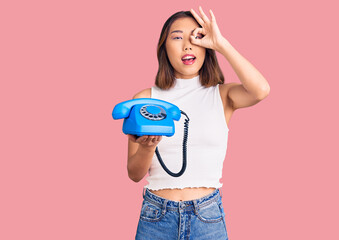 Young beautiful chinese girl holding vintage telephone smiling happy doing ok sign with hand on eye looking through fingers