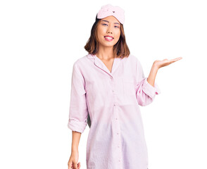 Young beautiful chinese girl wearing sleep mask and pajama smiling cheerful presenting and pointing with palm of hand looking at the camera.